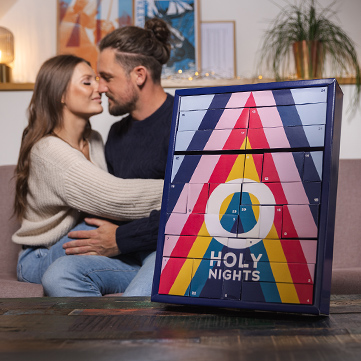 A couple sits on a sofa, hugging each other, with the ORION advent calendar in the foreground. The atmosphere is romantic and warm.