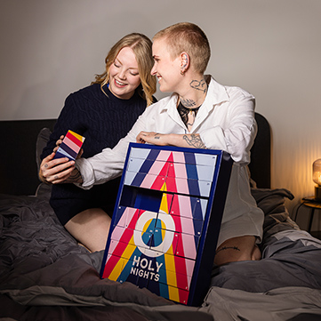 Zwei Frauen sitzen auf einem Bett und lachen, während sie eine Schublade des ORION Adventskalenders öffnen. Die Stimmung ist fröhlich und intim.